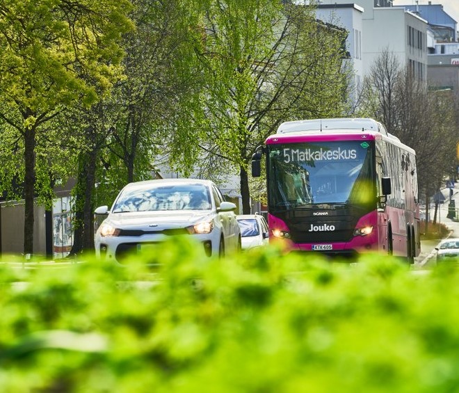 Auto ja linja-auto kadulla. Etualalla ja taustalla vihreää kasvillisuutta.